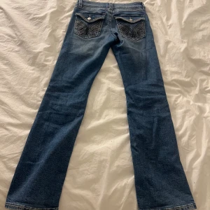 Blå bootcut jeans med broderade fickor - Säljer ett par blå jeans med bootcut-modell och snygga broderade bakfickor med knappar. Jeansen har klassisk femficksdesign och är tillverkade i jeansmaterial. Perfekta för dig som gillar en lite vidare passform nertill och coola detaljer.