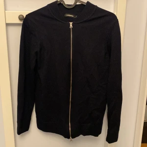 Mörkblå kofta från J.Lindeberg med dragkedja - Stilren Mörkblå kofta från J.Lindeberg i storlek S. Koftan har hel dragkedja framtill, ribbade muddar och är långärmad. Perfekt för lager-på-lager och enkel att matcha med olika outfits.