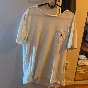 Vit t-shirt från Polo Ralph Lauren - Vit t-shirt från Polo Ralph Lauren med klassisk passform och korta ärmar. På bröstet finns en ficka med den ikoniska Polo-loggan broderad i mörkblått. Tillverkad i mjuk bomull, perfekt för en chill och clean stil.