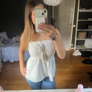 Vit smockad tubtopp från Zara - Superfin vit tubtopp från Zara med smockad överdel och volangkant upptill. Toppen har dekorativ knytning med tofsar och pärlor framtill. Tillverkad i mjukt bomullstyg som känns skönt mot huden. Perfekt för varma dagar och enkel att styla med jeans eller kjol.