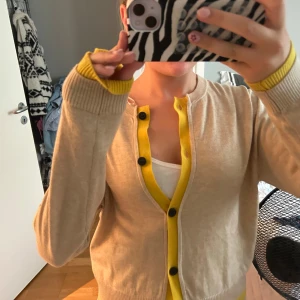 Beige kofta med gula detaljer - Superfin beige kofta med knallgula ribbade detaljer vid knäppning, ärmslut och nederkant. Svarta knappar framtill och rund halsringning. Perfekt för dig som vill ha en färgklick i garderoben! Mjuk och skön, passar till många outfits. Den är i strl M men sitter som S, endast andvänd 2 gånger ❣️