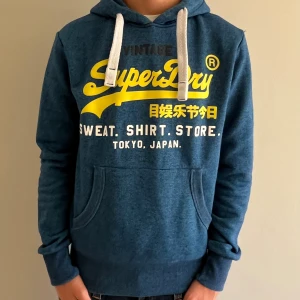 Blå hoodie från Superdry med tryck - Blå hoodie från Superdry med stort gult och vitt tryck på bröstet. Tröjan har huva med vita snören och en klassisk känguruficka framtill. Mjuk insida och ribbade muddar vid ärmslut och nederkant. Perfekt för en chill och sportig stil.