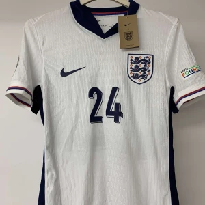 England fotbollströja Nike EM 24 - Snygg England fotbollströja från Nike med nummer 24 på bröstet och officiella EM 2024-patch på ärmen. Tröjan är vit med mörkblå detaljer och har korta ärmar med rödblå kant. Materialet är lätt och ventilerande polyester, perfekt för match eller träning.