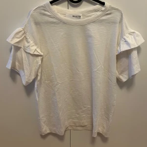 Vit t-shirt med volangärm från Selected Femme - Vit t-shirt från Selected Femme i mjuk bomull. Modellen har korta ärmar med volangdetalj som ger en trendig och söt look. Perfekt till jeans eller kjol för en fräsch och stilren outfit.
