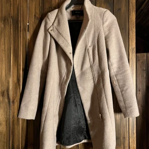 Beige kappa från Vero Moda - Säljer en stilren beige kappa från Vero Moda med snygg dragkedja framtill och klassisk krage. Den är lite  nopprig (se bild 2) och ser likadan ut på baksidan. Säljes då den är för liten och aldrig kommer till användning! 