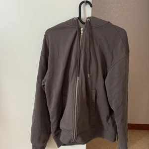 Brun hoodie med dragkedja från H&M - Mysig brun hoodie från H&M i loose fit-modell. Tröjan har huva med snörning, hel dragkedja framtill och två fickor. Tillverkad i mjuk bomullsblandning, perfekt för chill dagar. Passar dig som gillar en avslappnad och enkel stil.