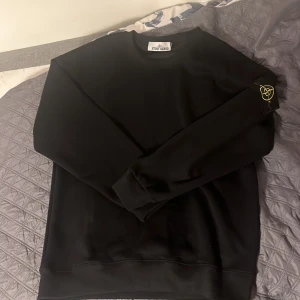Svart Stone Island sweatshirt M - Svart sweatshirt från Stone Island i storlek M. Klassisk rund hals och ribbade muddar. Ikonisk logopatch på vänster ärm. Tröjan har en clean och stilren look, perfekt för dig som gillar streetwear och vill ha något som sticker ut lite extra.