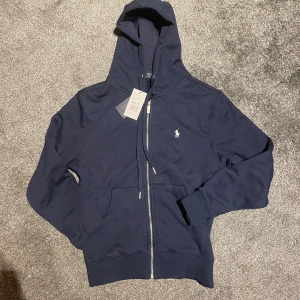 Marinblå hoodie från Polo Ralph Lauren - Snygg marinblå hoodie från Polo Ralph Lauren med dragkedja framtill, huva med snörning och klassisk logga broderad på bröstet. Tröjan har kängurufickor och ribbade muddar. Perfekt för en chill och stilren look.
