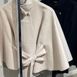 Beige cape med knyt och guldknapp - Intressekoll! Stilren beige cape från Missay med elegant knytband i midjan och dekorativa guldknappar vid halsen. Jackan har en vid, svepande form och hög krage som ger en sofistikerad look. Perfekt för dig som vill sticka ut med en unik och trendig ytterjacka. onesize.