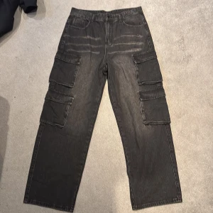 Svarta cargopants i denim - Säljer ett par svarta cargopants i denim med flera stora fickor på sidorna. Byxorna har raka ben och en avslappnad passform. Perfekta för dig som gillar streetwear och vill ha både stil och funktion. Klassisk femficksdesign framtill och två bakfickor.