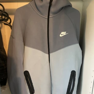Nike hoodie i ljusblått och grått - Nike hoodie i två nyanser av blått med dragkedja framtill och svarta detaljer vid fickorna. Tröjan har huva och Nike-logga på bröstet. Perfekt för en sportig och avslappnad stil.