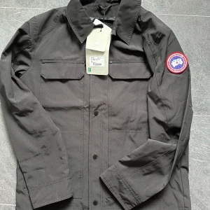 Svart overshirt från Canada Goose - Stilren svart overshirt från Canada Goose med klassisk krage, två bröstfickor med lock och ikoniskt rött märke på ärmen. Jackan har knäppning framtill och är tillverkad i slitstarkt material med slät finish. Perfekt för lager-på-lager och snygg till streetwear.
