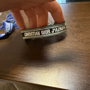 Christian Dior J'Adior armband svart/vitt - Snyggt armband från Christian Dior med texten 'J'Adior' broderad i vitt på svart bakgrund. Armbandet har justerbara tofsar i ändarna och är vävt i tyg med fina detaljer. Perfekt accessoar för att lyfta din outfit