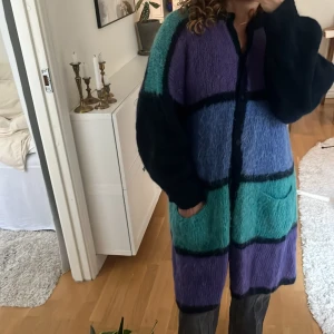 Färgglad lång ull kofta med knappar - Säljer en oversized lång kofta med stora blockrutor i lila, blått, grönt och svart. Koftan är stickad och har fluffig textur, två fickor framtill och knäppning med svarta knappar. Perfekt för dig som gillar färg och vill ha en mysig statement-piece.