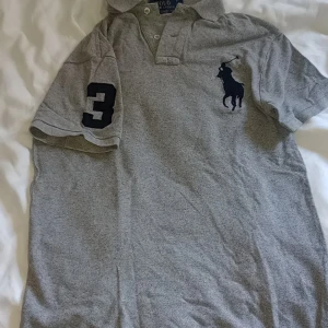 Grå Polo Ralph Lauren t-shirt S - Snygg grå t-shirt från Polo Ralph Lauren i slim fit med korta ärmar och klassisk krage. Stor svart polospelare broderad på bröstet och en stor svart siffra 3 på ena ärmen. Tillverkad i mjuk bomull, perfekt för en chill och stilren look.