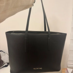 Svart axelväska från Valentino - Stilren svart axelväska från Valentino i slätt skinn med guldfärgad dragkedja och diskret logga framtill. Väskan har långa handtag, rymligt huvudfack och innerficka med dragkedja. Perfekt för dig som vill ha en klassisk och snygg väska med plats för allt du behöver.