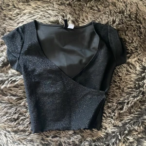 Glittrig svart omlottopp från H&M Divided - Snygg svart omlottopp från H&M Divided i storlek XS. Toppen har glittrigt tyg, korta ärmar och djup v-ringning. Perfekt för dig som vill sticka ut med lite extra glitter. Materialet känns mjukt och följsamt mot huden.