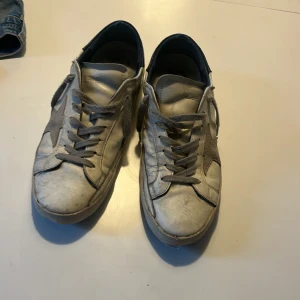 Golden Goose Superstar sneakers vit/grå - Golden Goose Superstar sneakers i vitt skinn med grå mockadetaljer och stjärna på sidan. Skorna har grå snörning, blå hälkappa och GGDB/SSTAR-tryck på sidan. Klassisk platt sula och rund tå. Perfekt för dig som gillar en avslappnad och trendig look. Säljer billigt då det är väldigt slitna kan tänka mig gå ner i pris vid övriga frågor är bara att höra av sig