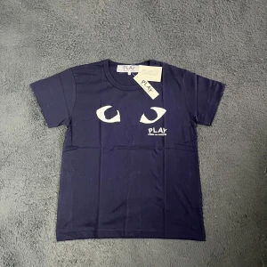Comme des Garçons t-shirt - Helt ny Mörkblå t-shirt från Comme des Garçons Play med ikoniskt ögontryck och logga på bröstet. Klassisk rund hals, korta ärmar och tillverkad i mjuk bomull. Perfekt för dig som gillar streetwear och vill sticka ut med en stilren men unik look. Axelbredd 43cm Längd 64cm. JAG KAN EJ SKICKA EN BILD NÄR JAG BÄR DEN DÅ JAG EJ KAN HA STORLEKEN. 