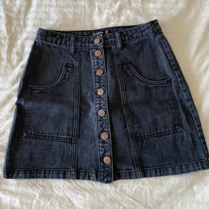 Svart jeanskjol med knappar - Snygg svart jeanskjol med stora fickor framtill och dekorativa knappar längs hela framsidan. 