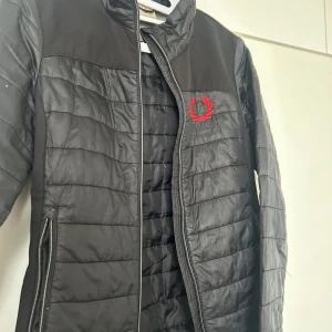 Svart pufferjacka - Svart pufferjacka från Frinter med broderad röd lagerkrans på bröstet. Jackan har quiltad yta, dragkedja framtill och sidofickor med dragkedja. Insidan är fodrad med mjuk fleece för extra värme. Snygg och sportig look, perfekt för kyliga dagar.