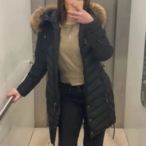 Svart lång dunjacka med päls - Säljer en svart lång hollies pufferjacka med snygga horisontella sömmar och dragkedja framtill. Jackan har hög krage och praktiska fickor med dragkedja. Perfekt för kalla dagar, äkta päls 