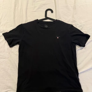 Svart t-shirt från GANT - En stilren svart t-shirt från GANT med liten broderad logga på bröstet. Klassisk rund halsringning och korta ärmar. Perfekt basplagg i garderoben och tillverkad i mjuk bomull för skön känsla.