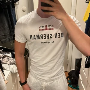 Vit Ben Sherman t-shirt med tryck - Säljer en vit t-shirt från Ben Sherman med brittisk flagga och svart text på bröstet. Klassisk passform och rund hals. Perfekt för dig som gillar stilrena och ikoniska plagg med en touch av brittisk vibe.