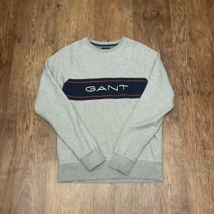 Grå sweatshirt från GANT med logga - Grå sweatshirt från GANT i storlek M med en mörkblå panel och vit logotyp framtill. Tröjan har rund hals, ribbade muddar och lång ärm. Perfekt för dig som gillar klassisk och enkel stil med en touch av street.