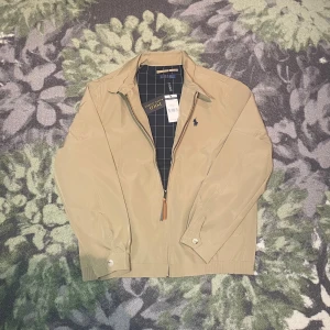 Beige vindjacka från Polo Ralph Lauren - Snygg beige vindjacka från Polo Ralph Lauren med klassisk krage och dragkedja framtill. Jackan har mörkblått foder med vitt rutmönster och ikonisk broderad logga på bröstet. Tillverkad i bomull och polyester, perfekt för vår och höst. Storlek S