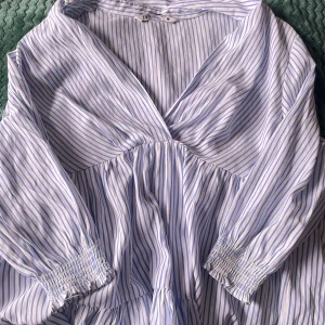 Randig omlottklänning från Zara - Superfin kort omlottklänning från Zara i vitt och ljusblått med smala ränder. Klänningen har v-ringning, långa ärmar med smock vid handlederna och volangig kjol. Perfekt för sommarens alla tillfällen!