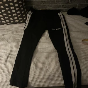 Palm Angels svarta trackpants med vita ränder - Svarta trackpants från Palm Angels med vita sidoränder och logga tryckt på låret. Byxorna har en slim passform och är perfekta för en streetwear-look. Materialet känns mjukt och stretchigt, vilket gör dem riktigt sköna att bära.