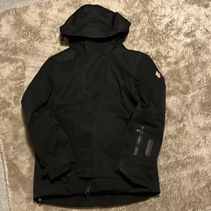 Svart Moncler skiijacket - Svart vindjacka från Moncler med huva och diskret logga på ärmen. Jackan har dragkedja, flera fickor och är tillverkad i ett slitstarkt syntetmaterial. Insidan är quiltad för extra komfort och värme. Perfekt för dig som vill ha en stilren och funktionell jacka.
