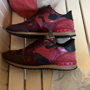 Valentino Garavani Rockrunner sneakers - Säljer ett par Valentino Garavani Rockrunner sneakers i röd och vinröd färg med svart och mörkrött camouflagemönster. Skorna har detaljer i mocka och skinn, klassisk snörning och ikoniska nitar på hälen. Snygg och unik design som sticker ut.