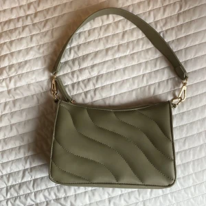 Olivgrön quiltad axelväska - Snygg olivgrön axelväska med vågformad quiltad design och bred axelrem. Väskan har guldfärgade metalldetaljer och dragkedja upptill. Perfekt storlek för det viktigaste och ger en trendig touch till din outfit.
