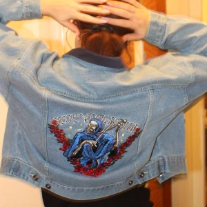 Jeansjacka HUF med broderad rygg - Säljer en blå jeansjacka från HUF med mörk krage och stora bröstfickor. På ryggen finns ett stort broderi med dödskalle i blå kåpa omgiven av röda rosor och texten 'HUF Worldwide'. 