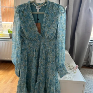 Kortklänning från Aware - Supersöt kortklänning från Aware i ljusblått med blommigt mönster. Klänningen har v-ringning, långa puffärmar med knappar vid ärmslut och markerad midja. Perfekt för vår och sommar, i ett lätt och luftigt material. Oanvänd 