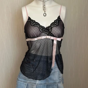 Svart mesh linnetopp med spets och rosett - Säljer en svart mesh linnetopp med tunna rosa axelband, spetskant vid bysten och en söt rosa rosett under bysten. Toppen är genomskinlig med små prickar i tyget och har en romantisk vibe. Perfekt att styla lager på lager eller som statement-piece.