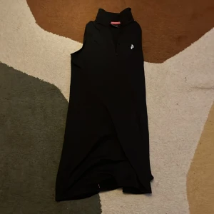 Svart träningsklänning Peak Performance - Sportig svart träningsklänning från Peak Performance med krage och knappslå framtill. Ärmlös design och stilren look, perfekt för dig som gillar att vara aktiv och ändå se snygg ut. Diskret logga på bröstet och mjukt, funktionellt material.