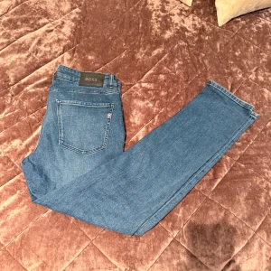 Blå slim-fit jeans från BOSS - Snygga blå jeans från BOSS i slim fit. Jeansen har en stilren look med diskret BOSS-logga på bakfickan och svart patch i midjan. Perfekta för dig som gillar enkel och tidlös stil. Dessa jeans är i riktigt bra skick, har används några gånger. 