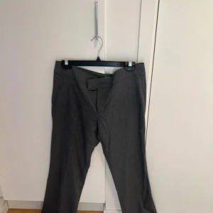 Grå kostymbyxor med bred linning - Snygga grå kostymbyxor med bred linning och dold knäppning framtill. Byxorna har raka ben och en klassisk look som passar perfekt till en dressad outfit. Materialet är mjukt och ger en stilren känsla. Lite noppriga på insidan av byxbenet