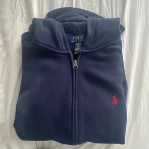 Marinblå hoodie från Polo Ralph Lauren - Marinblå hoodie från Polo Ralph Lauren med dragkedja och huva. Klassisk röd broderad logga på bröstet och kängurufickor framtill. Tillverkad i mjuk bomullsmix, perfekt för chill dagar eller när du vill ha en clean look.