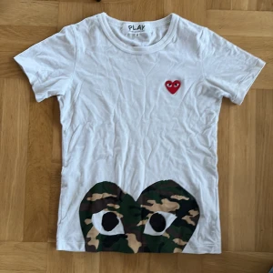 Vit t-shirt från Comme des Garçons Play med camo-detalj - Vit t-shirt med det klassiska röda hjärtat på bröstet och en stor camo-mönstrad hjärtdetalj med ögon längst ner. Enkel och stilren design som sticker ut med coola detaljer.