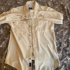 Beige skjorta från M.G. Star - Stilren beige skjorta från M.G. Star med klassisk krage, två bröstfickor med lock och vita knappar. Skjortan har lång ärm och är tillverkad i ett strukturerat tyg som känns mjukt och bekvämt. Perfekt för dig som gillar en clean och enkel look.