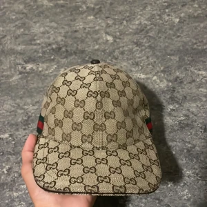 Gucci keps med GG-mönster och rödgrön detalj - Säljer en beige keps från Gucci med klassiskt GG-monogram i brunt över hela kepsen. På sidan finns den ikoniska rödgröna randen. Justerbar rem baktill och insida med svart foder. Perfekt för dig som vill sticka ut med lyxig streetstyle.pris kan diskuteras.