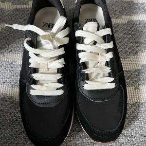 Svarta sneakers från Zara med vit sula - Svarta sneakers från Zara med vit platt sula och grovt mönstrad undersida i ljusbrunt. Skorna har vita snören, rund tå och detaljer i mocka och syntet. Snygg kontrast mellan svart och vitt, perfekt för dig som gillar stilrena och sportiga vibes.