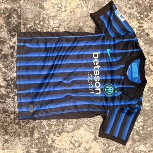 Fotbolls tröja  - Snygg Inter Milan fotbollströja med Lautaro och nummer 10 på ryggen. Svart och blått randigt mönster, klassisk Nike-logga och sponsortryck på bröstet. Kortärmad modell i lätt och ventilerande polyester, perfekt för match eller träning.