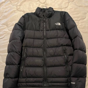 Svart dunjacka från The North Face - Säljer en svart dunjacka från The North Face med klassisk puffer-design. Jackan har hög krage, två dragkedjefickor och logga på bröstet och ryggen. Perfekt för kalla dagar och riktigt snygg streetstil.