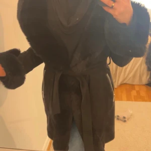 Svart pälsjacka med bälte och dragkedjor - Snygg svart pälsjacka med bred pälskrage och pälskanter vid ärmsluten. Jackan har knytskärp i midjan och silverfärgade dragkedjor på sidorna. Perfekt för kalla dagar när du vill se extra lyxig ut.