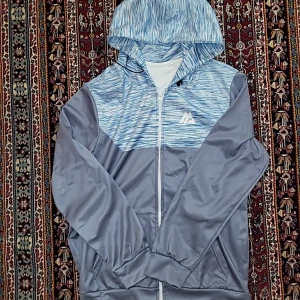 Blå och grå Montirex vindjacka med huva - Snygg vindjacka från Montirex i blått och grått med mönstrad överdel och huva. Jackan har dragkedja framtill, långa ärmar och är tillverkad i polyester. Perfekt för dig som gillar sportig stil och vill ha något lätt och smidigt.